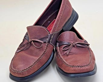 May include: A pair of brown leather loafers with a bow detail on the vamp. The shoes have a textured finish, white stitching, and black soles. The shoes are a size 8.