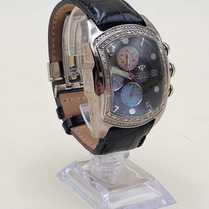 May include: An Aqua Master chronograph watch with a black leather strap, a silver-tone rectangular case featuring a diamond-encrusted bezel, a dark dial with three sub-dials, a date window at the three o'clock position, and silver-tone markers.