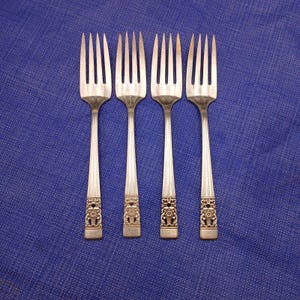 May include: Four silver forks with ornate gold designs on the handles. The forks are arranged in a row on a blue background.