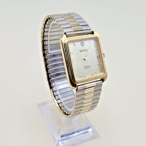May include: A Benrus quartz wristwatch with a rectangular gold-tone case and a light-coloured dial featuring a single diamond marker at the twelve o'clock position. The watch has a two-tone gold and silver expandable metal link bracelet.