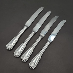 Puede incluir: Cuatro cuchillos plateados con un diseño decorativo de concha en los mangos. Las hojas son lisas y los mangos tienen un patrón acanalado. Los cuchillos están dispuestos en diagonal sobre un fondo oscuro.