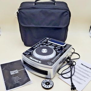 May include: A silver and black Numark NDX 400 DJ media player featuring a central jog wheel, digital display, and front-loading disc slot. The set includes a black carrying bag, a power cord, a small circular dial, and two printed instruction manuals.