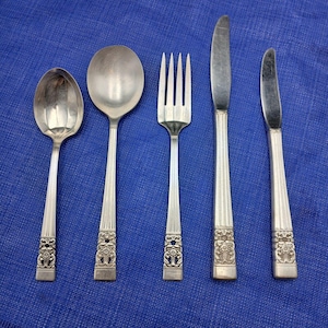 Könnte beinhalten: Fünf Besteckteile, darunter eine Gabel, ein Buttermesser, ein Essmesser und zwei Löffel, alle mit verzierten Griffen. Das Besteck ist silberfarben und hat eine polierte Oberfläche.