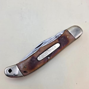 May include: A closed folding pocket knife with a brown, mottled handle and silver-toned metal bolsters. A silver metal shield is embedded in the handle with the text OLD TIMER engraved in black. The knife features a small hole at one end.