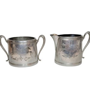 May include: A silver-toned metal sugar bowl with two curved handles and a matching creamer pitcher with a single handle and a pouring spout. Both pieces feature horizontal line detailing at the top and bottom rims and an engraved floral motif.