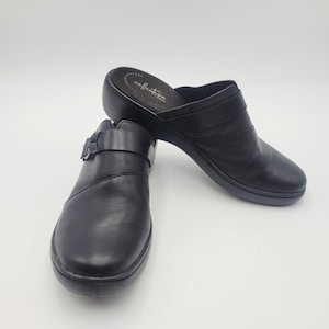 May include: A pair of black leather clogs with a buckle closure and a rounded toe. The shoes have a low heel and a cushioned insole. The word "collection" is printed on the insole. The clogs are set against a white background.