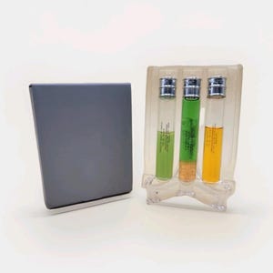 May include: Three glass vials with silver caps, each containing a different colored liquid: green, yellow, and orange. The vials are in a clear plastic holder next to a gray rectangular object. The vials have text on them.
