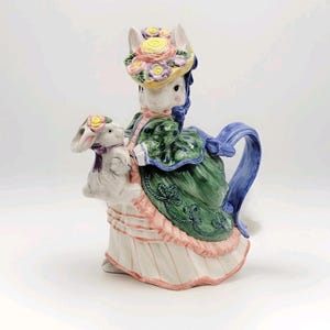May include: A ceramic teapot shaped like a woman with bunny ears, wearing a green dress and a floral hat. The teapot is holding a white bunny with a purple bow. The handle and spout are blue.