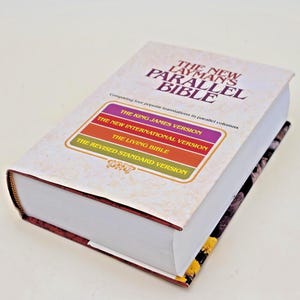 May include: A thick hardcover book titled The New Layman's Parallel Bible with a white cover. The text lists four translations: The King James Version, The New International Version, The Living Bible, and The Revised Standard Version in colored boxes.