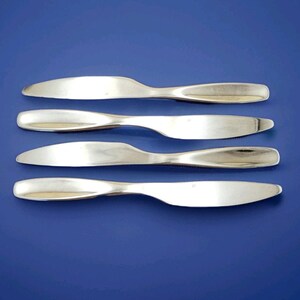 May include: Four silver butter knives arranged horizontally on a solid blue background. The knives have a curved blade and a rounded handle. The blades are reflective, and the handles are smooth.