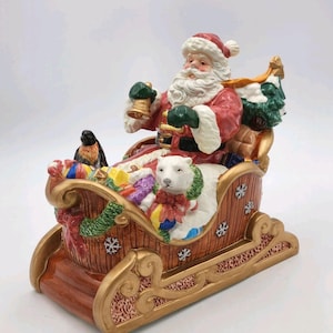 Puede incluir: Una figurita de cerámica de Santa Claus montando en un trineo lleno de regalos. Santa lleva un traje rojo con barba blanca y un gorro verde. El trineo es marrón con detalles dorados y está lleno de regalos coloridos, un oso polar y un pájaro.