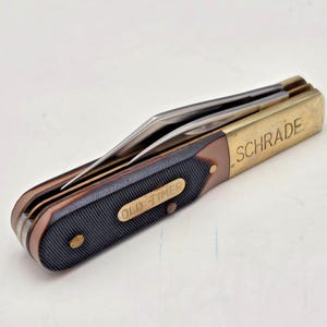 Könnte beinhalten: Ein klappbares Taschenmesser mit geschlossenem Griff und texturierten dunkelbraunen Griffschalen. Eine Messingbacke ist mit dem Schriftzug SCHRADE graviert, und eine Messingeinlage auf dem Griff zeigt den Text OLD TIMER. Metallklingen sind sichtbar.