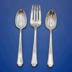 May include: Three pieces of silver cutlery, a fork and two spoons, are displayed against a blue backdrop. The cutlery has decorative patterns on the handles. The fork has four prongs, and the spoons have oval bowls.