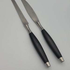 May include: Two silver knives with black handles. The knife on the left has a serrated edge. The knife on the right has a smooth edge.