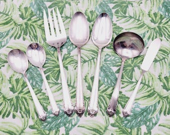 May include: A set of seven silver-plated utensils, including a fork, a spoon, a ladle, a butter knife, and three spoons. The utensils are arranged on a green and white patterned tablecloth.