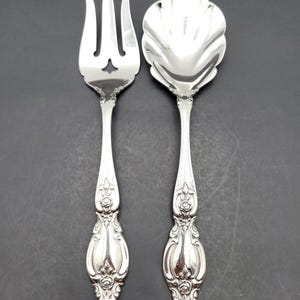 Könnte beinhalten: Ein Set aus zwei versilberten Servierbesteck, eine Gabel und ein Löffel. Die Gabel hat einen dekorativen Griff mit einem floralen Design. Der Löffel hat eine muschelförmige Schale und einen dekorativen Griff mit einem floralen Design.