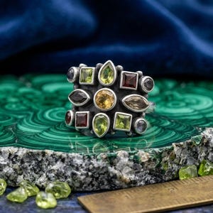 Op de afbeelding: Een ring van sterling zilver met een centrale ronde gele edelsteen omringd door diverse stenen in verschillende vormen, waaronder groene peridoot, rode granaat en bruine kwarts. Een metalen liniaal toont een breedte van circa 2,5 cm.