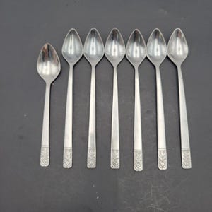 Pode incluir: Conjunto de sete colheres de prata. As colheres têm uma tigela em forma de gota e um cabo longo e fino. O cabo apresenta um desenho floral na base. As colheres estão dispostas sobre uma superfície escura, voltadas para cima.