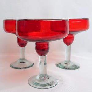 May include: Three red glass margarita glasses with clear stems and bases. The glasses are arranged in a row on a white surface.