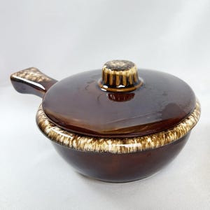 May include: A brown ceramic casserole dish with a lid and handle. The dish has a glossy finish with a lighter brown and white trim around the rim and on the handle. The lid has a small, textured knob.