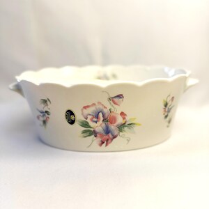 May include: Off-white ceramic serving bowl with a scalloped rim and two small handles. The bowl is decorated with floral designs in shades of pink, purple, and green. A small emblem is visible on the side.