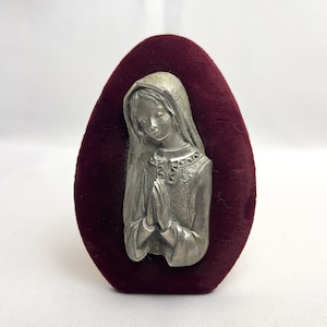 May include: A silver-colored religious plaque depicting a woman with clasped hands, set against a deep burgundy velvet oval. The woman is wearing a head covering and a detailed gown. The velvet has a soft, plush texture.