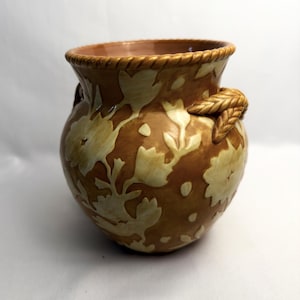 May include: A round ceramic vase featuring a glossy brown glaze with a cream-coloured floral pattern. The rim has a textured rope-like design, and two sculpted, braided handles are attached to the sides of the vessel, adding dimension.