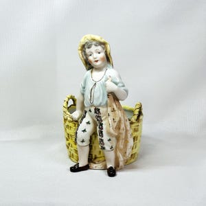 May include: A ceramic figurine of a young person in a light blue shirt, patterned pants, and a straw hat, holding a tan sack. The figure is posed next to a yellow basket with a woven design. Black shoes complete the outfit.