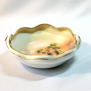 May include: A small, white ceramic bowl with a scalloped rim and gold trim. The interior features a painted landscape scene with green trees and a peach-colored sky. A decorative piece.