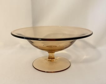 May include: A translucent amber-colored glass bowl with a wide, shallow basin and a smooth, rounded rim. The bowl is supported by a short, central pedestal stem that connects to a circular, flat base, creating a balanced, elevated silhouette.