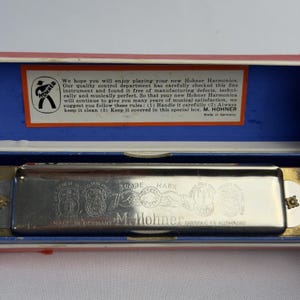 May include: A silver-toned metal Hohner harmonica rests inside a red, white, and blue case. The harmonica features engraved text including M. Hohner, Trade Mark, and Made in Germany. The case lid contains a printed note about the instrument.