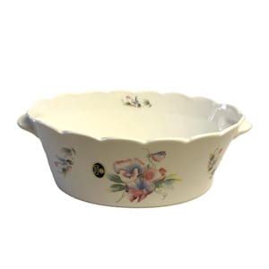 May include: Cream-colored ceramic serving bowl with a scalloped rim and two small handles. The bowl is decorated with a floral pattern in shades of pink, blue, and green. A small emblem is visible on the side of the bowl.