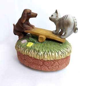 May include: A ceramic figurine featuring a brown dachshund and a grey tabby cat on a seesaw. The base is shaped like a grassy mound with small yellow and pink flowers, resting on a textured, reddish-brown stone-patterned foundation.