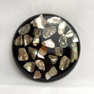 May include: A round, black decorative object inlaid with iridescent, irregularly shaped pieces. The pieces have a pearlescent sheen with hints of brown, silver, and gold. The object is set against a white background.