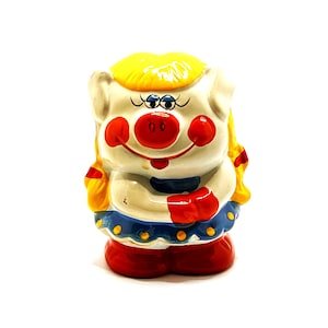 May include: A ceramic piggy bank shaped like a pig with yellow hair, red shoes, and a blue and yellow dress. The pig has a white face with red cheeks and a red nose.