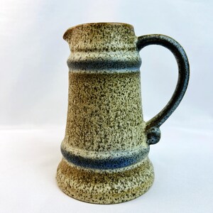 May include: A ceramic pitcher featuring a speckled beige and brown finish with two horizontal blue bands encircling the body. The vessel has a flared base, a cylindrical shape, a small pouring spout, and a curved handle attached to the side.