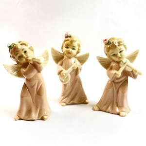 May include: Three ceramic angel figurines in a soft peach color. Each angel has blonde hair, delicate wings, and a small floral crown. One plays a flute, another a banjo, and the third a violin. A charming decorative set.