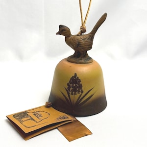May include: A ceramic bell featuring a brown bird figurine on top and a gradient finish transitioning from tan to dark brown. A dark brown floral illustration is centered on the bell. A tan paper tag with the text The Legend of the Windbells is attached.