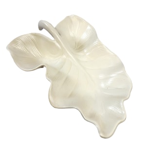 May include: A cream-colored ceramic dish shaped like a large tropical leaf. The surface features embossed, radiating vein patterns that extend from a central stem. The edges are scalloped and follow the organic, asymmetrical contour of a leaf.