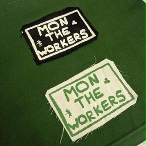 Op de afbeelding: Een donkergroene stoffen achtergrond met twee rechthoekige patches. De bovenste patch is zwart met witte geborduurde tekst MON THE WORKERS. De onderste patch is wit met groene gedrukte tekst MON THE WORKERS.