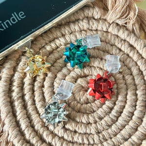 May include: Four different colored bow-shaped charms with clear plastic clasps. The charms are gold, green, red, and silver. The charms are on a woven brown surface.