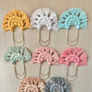 May include: Nine colorful crocheted bookmarks with gold metal clips. The bookmarks are in the shape of a half circle with fringe around the edges. The colors are yellow, white, light blue, light green, pink, peach, gray, and two shades of pink.
