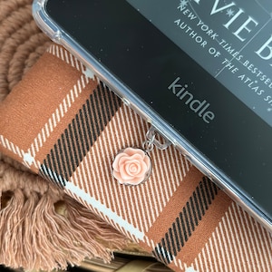 May include: A close-up of a Kindle e-reader with a clear case and a pink flower charm attached. The e-reader is on a brown and black plaid surface.