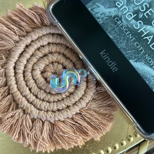 May include: A holographic charm in the shape of the letter "S" on a brown woven coaster with fringe. The charm is attached to a black Kindle e-reader with a case. The e-reader is displaying the text "House of  and Shad A Crescent City Novel kindle".