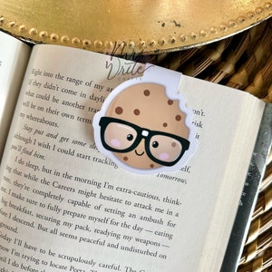 May include: A brown cookie bookmark with a white outline and a pair of black glasses. The cookie has a smiling face with pink cheeks. The bookmark is placed on a book with text that reads "Right into the range of my arm. If they didn't come in daylight, what could be another trap?"