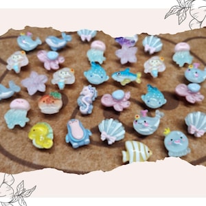 May include: A collection of miniature ocean-themed resin charms. The charms include various sea creatures like whales, fish, seahorses, and starfish in pastel colors. They are arranged on a brown surface, perfect for crafting or jewelry making.