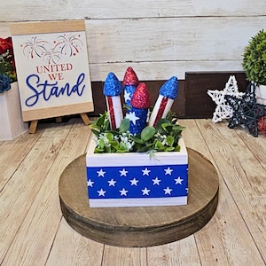 May include: A patriotic centerpiece featuring a white box with a blue star-patterned ribbon. The box holds faux greenery and decorative firework-shaped ornaments with red, white, and blue glitter. A framed American flag and a sign that reads "United We Stand" are in the background.