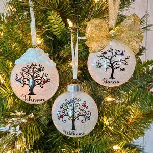 May include: Three white glass Christmas ornaments with a glittery finish. Each ornament features a black silhouette of a tree with colorful gems and a name at the bottom: Johnson, Garcia, and Williams.