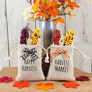 May include: Two small burlap bags with drawstrings, filled with faux corn cobs. One bag is labeled "Happy Harvest" and the other is labeled "Harvest Market". Both bags have a black and white checkered ribbon tied around them.