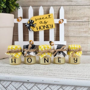 May include: Five small glass jars with yellow and white gingham lids and twine tied around the tops. The jars are filled with a clear liquid and labeled with the letters "H", "O", "N", "E", and "Y". The jars are sitting on a white surface in front of a white picket fence with a sign that reads "sweet as honey".